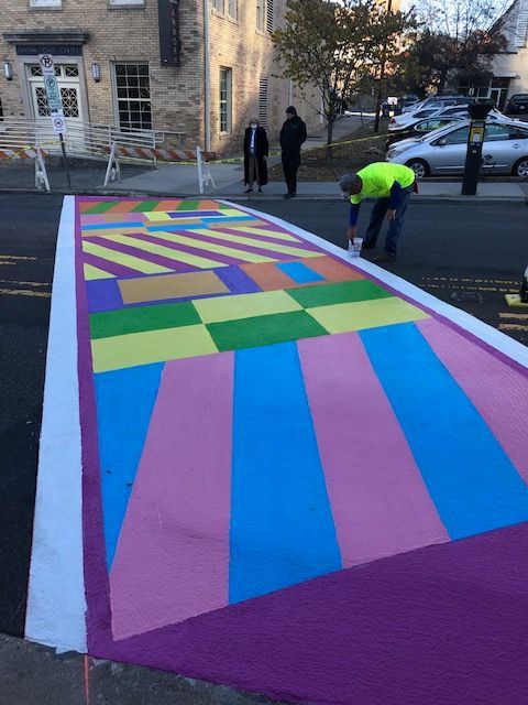 Person painting a colorful geometric street mural on a road beside parked cars and a stone building