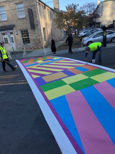 Workers painting a colorful striped crosswalk on a street beside stone buildings.