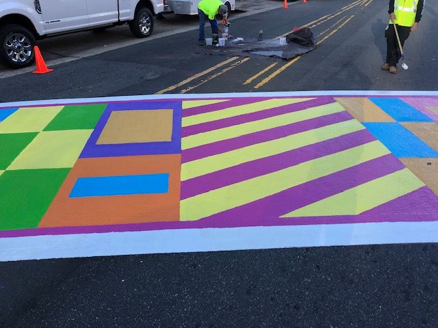 Colorful painted crosswalk with diagonal stripes on a street, with workers and traffic cones nearby.