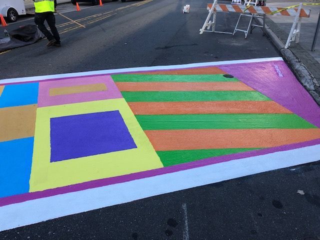 Colorful striped road marking painted on asphalt near orange barricades and a worker in a high-visibility vest.