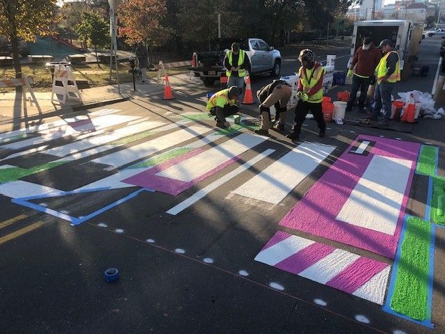 Workers painting a colorful street mural with pink, blue, black, and green geometric shapes
