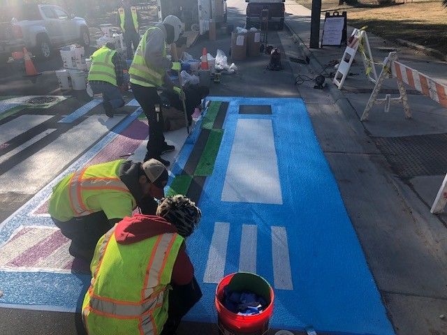 Workers painting a blue road mural on a street, wearing high-visibility vests and kneeling by paint buckets.