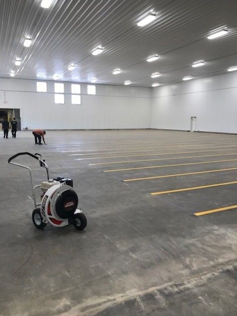 Large empty warehouse with overhead lights, a floor-scrubbing machine in the foreground, and two people near the back wall