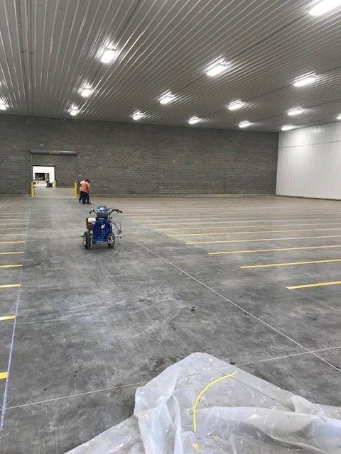 Large empty warehouse with gray concrete floor, overhead lights, and two workers using equipment near the back wall.