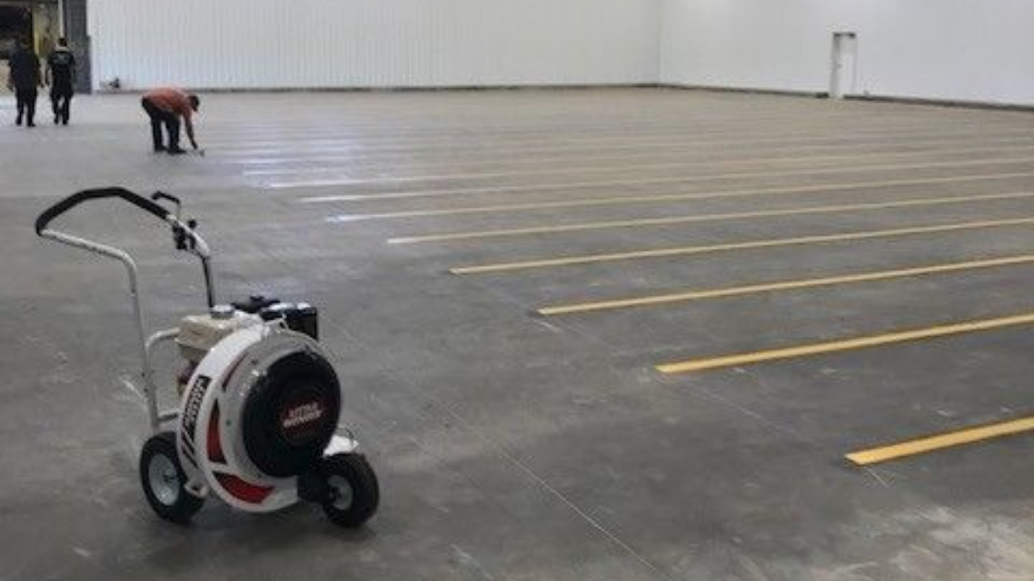 White floor-marking machine in a large empty warehouse, with a person working in the distance.