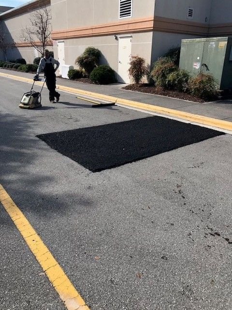 Worker pressure-washing a dark patch on an asphalt lot beside a beige building