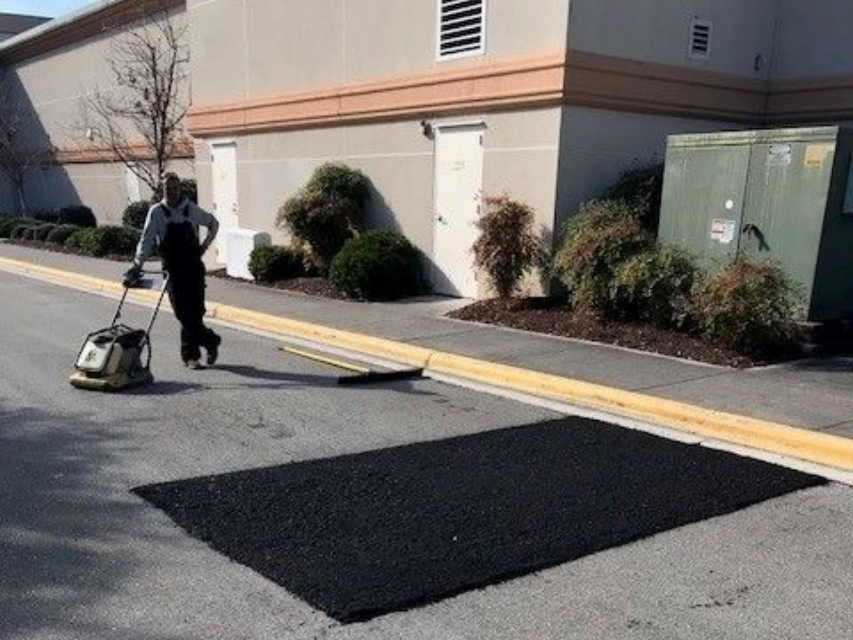 Worker sealing a sidewalk crack beside a building, with fresh black asphalt patch on the pavement in Fayetteville NC