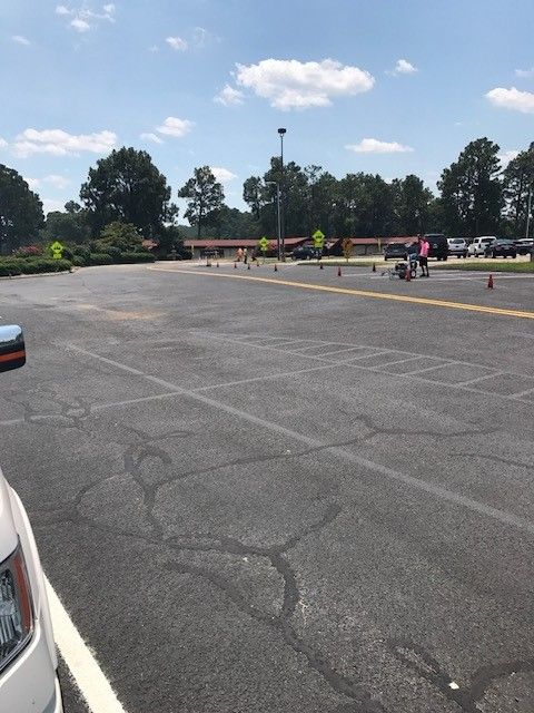 Wide parking lot with a car on the left, cones, and a few people riding scooters in the distance under a sunny sky