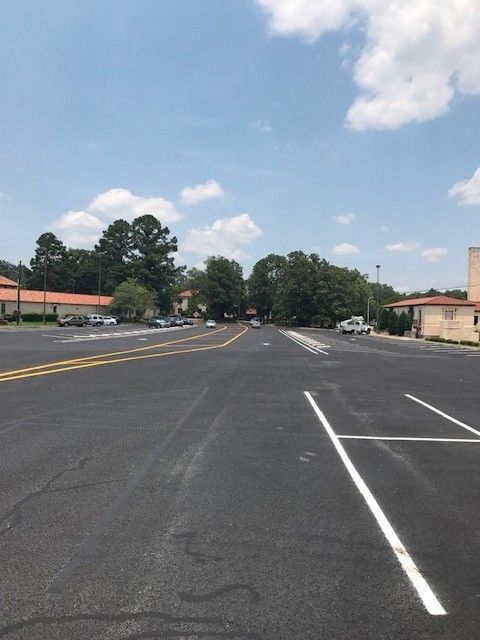 Empty asphalt parking lot with white lines under a partly cloudy sky, bordered by trees and low buildings