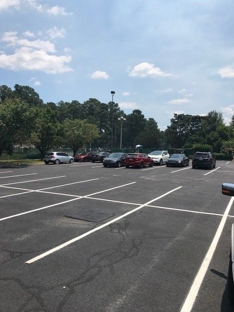 Mostly empty parking lot with a few parked cars, trees, and a partly cloudy sky.