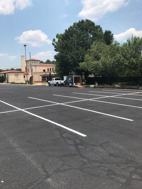 Empty parking lot with white lines, a few parked vehicles, and a building beside a large tree under a cloudy sky