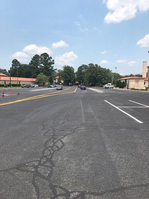 Wide cracked parking lot with a few cars, trees, and low buildings under a partly cloudy sky