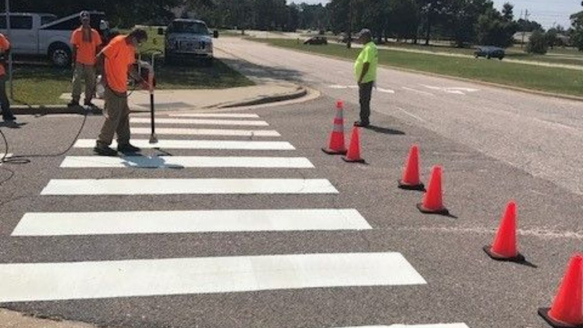 Road workers painting white crosswalk stripes beside orange cones on a street in Fort Bragg, NC.