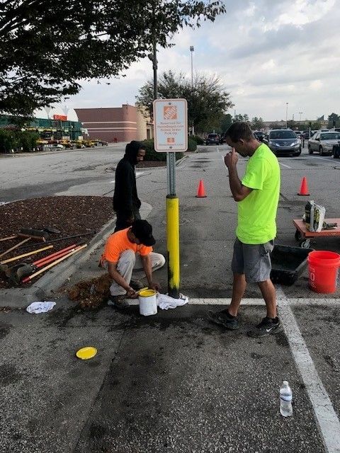 People painting a yellow curb line in a parking lot, with cones and tools nearby.