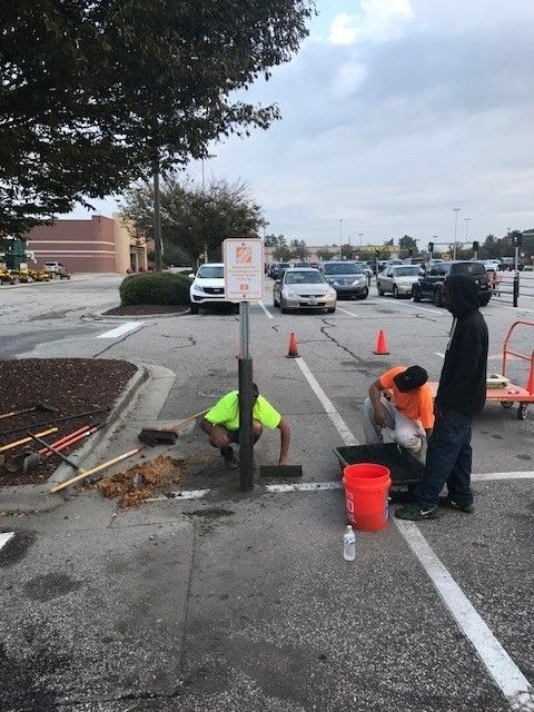 Road workers repairing a pothole on a parking lot lane, with cones, tools, and a bucket.