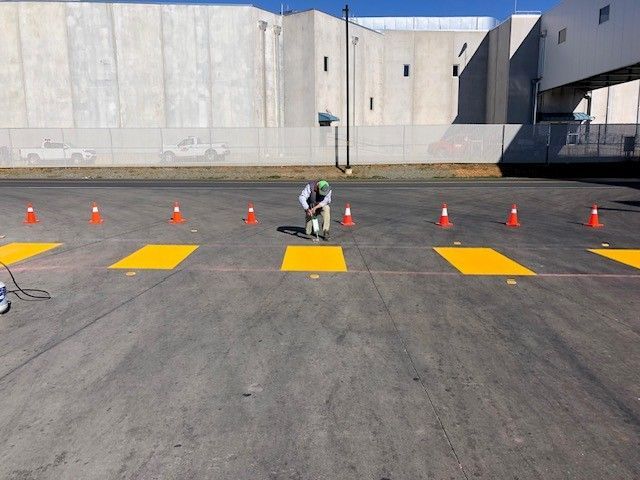 Person painting yellow stripes on asphalt with orange cones in a parking lot beside a large concrete building