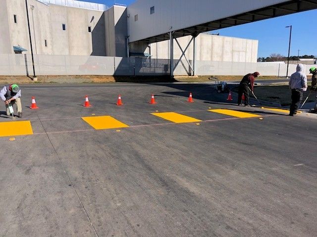 Workers marking an asphalt lot with orange cones and yellow painted rectangles beside a concrete building.