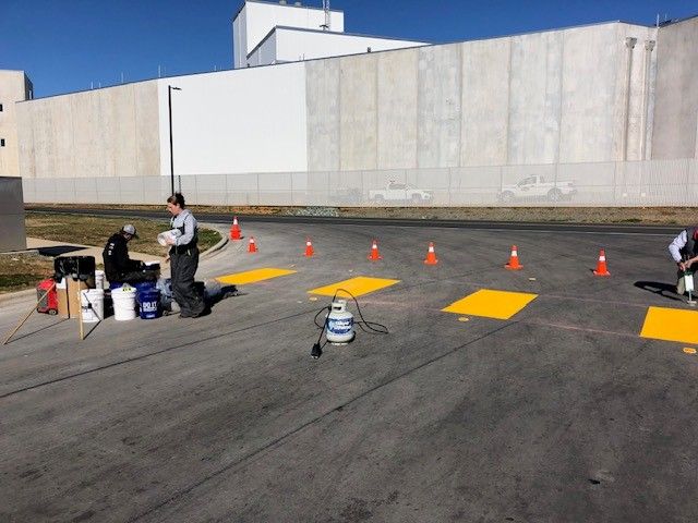 People and equipment on a paved lot with cones and yellow markers beside a large white building