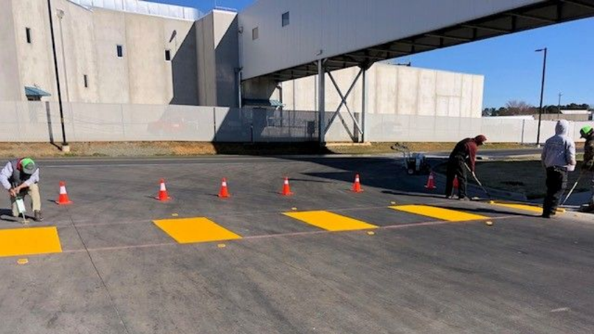 Workers set up orange cones and yellow markings on a paved lot beside a building and skybridge.