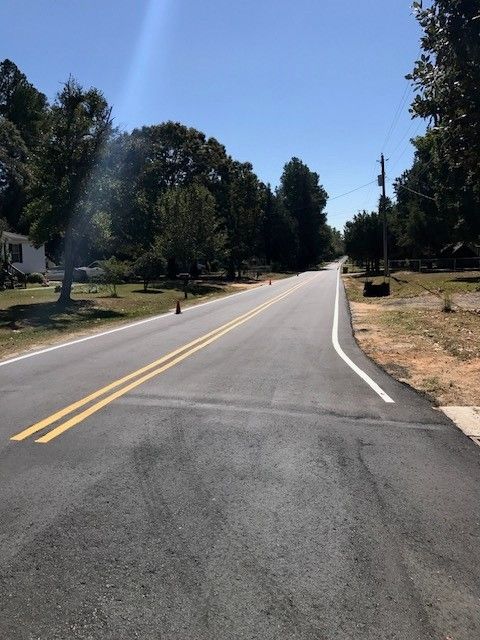 Quiet rural road curving uphill, lined with trees and houses under a clear blue sky.