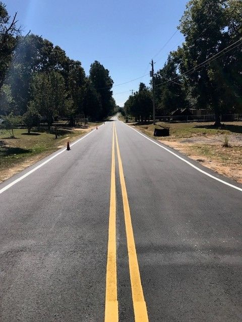 Empty two-lane road with double yellow lines through a tree-lined rural area under a clear blue sky
