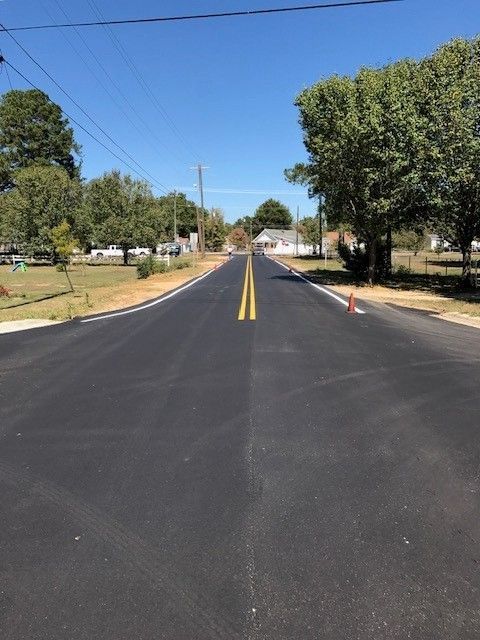 Freshly paved suburban road with double yellow lines, trees, and orange traffic cones under a clear blue sky