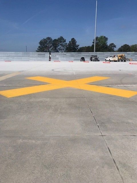 Yellow X painted on an empty concrete rooftop with a few distant trees and parked vehicles under a blue sky
