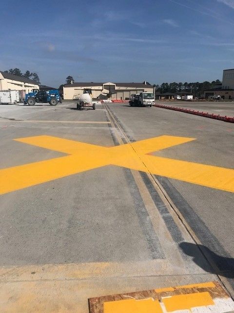 Yellow X painted on a concrete tarmac with buildings and equipment in the background.