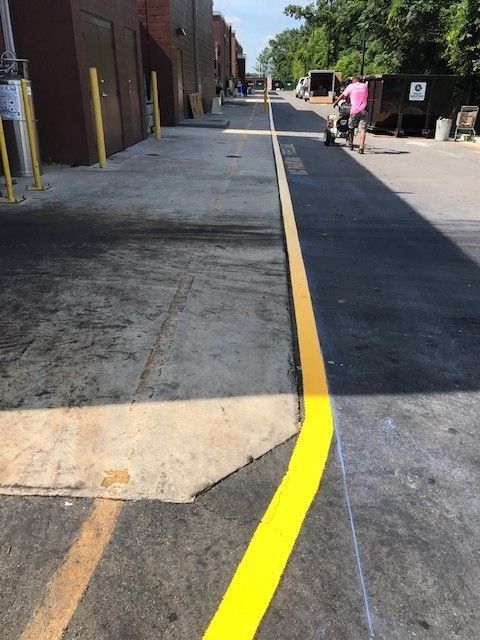 Yellow painted curb line along a sidewalk beside an industrial building and parked vehicles