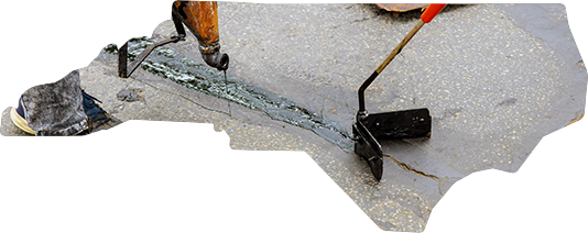 A worker uses a hot crack-sealing tool to apply sealant into a crack on a grey asphalt surface.