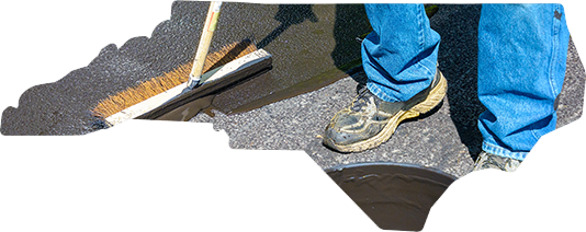 A worker in blue jeans uses a stiff-bristled brush to apply black asphalt sealer to a driveway surface.