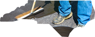 A person in blue jeans uses a push broom to spread black sealant over asphalt, shaped like a map of North Carolina.