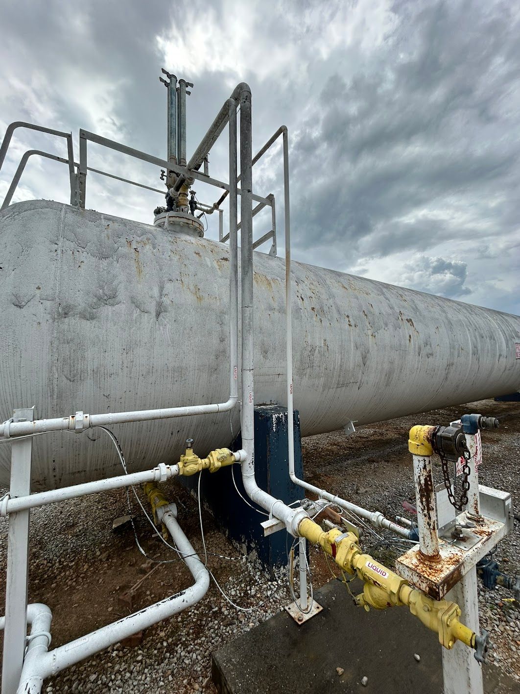 Large white industrial tank with piping, ladders, and valves, against a cloudy sky.
