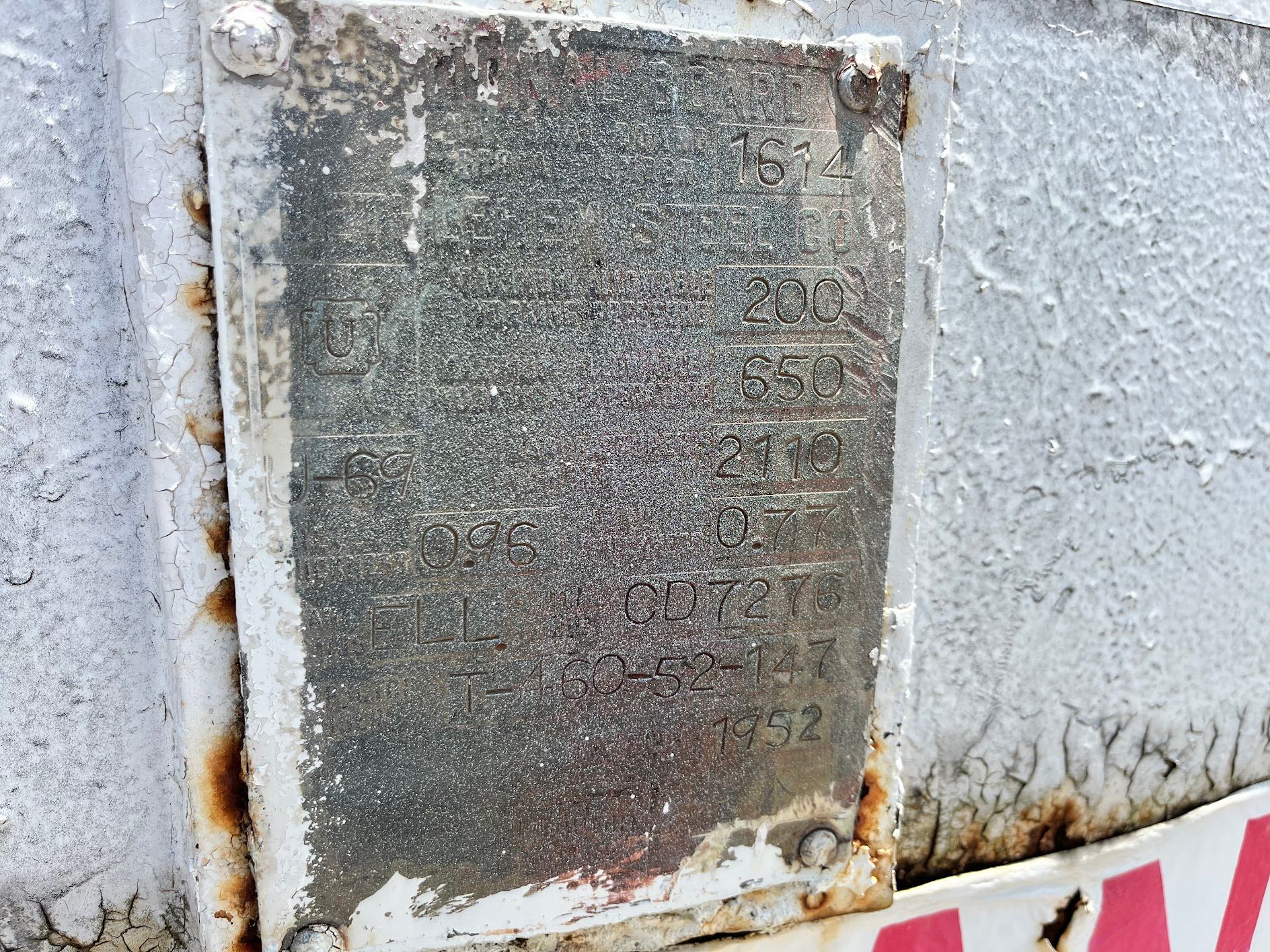 Faded metal plate with text and numbers, attached to a white and rusty surface.