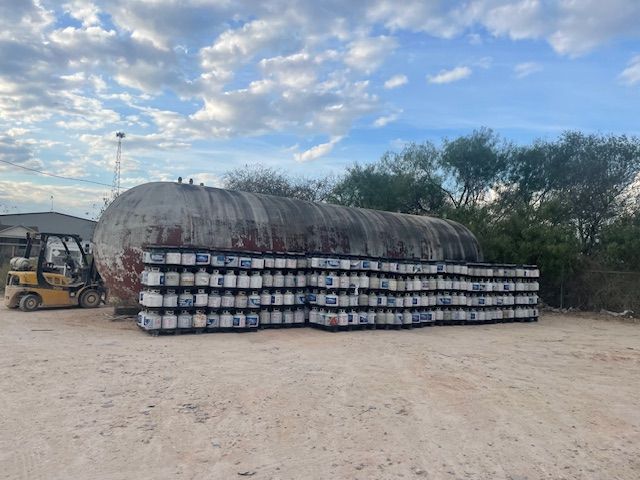 Forklift next to a large tank and stacks of white containers on pallets outdoors under a cloudy sky.