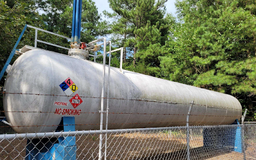 Large, horizontal white propane tank with safety placards, set against trees and a chain-link fence.