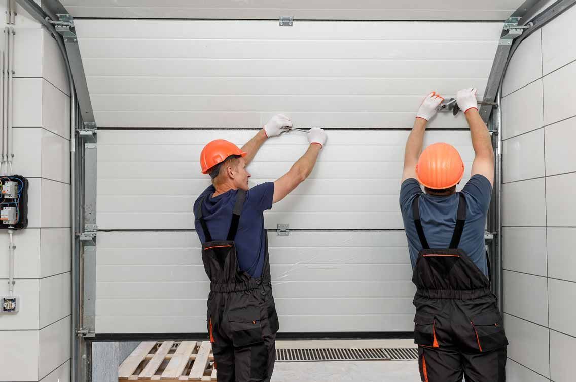 Two workers in orange helmets installing a garage door in a white industrial space