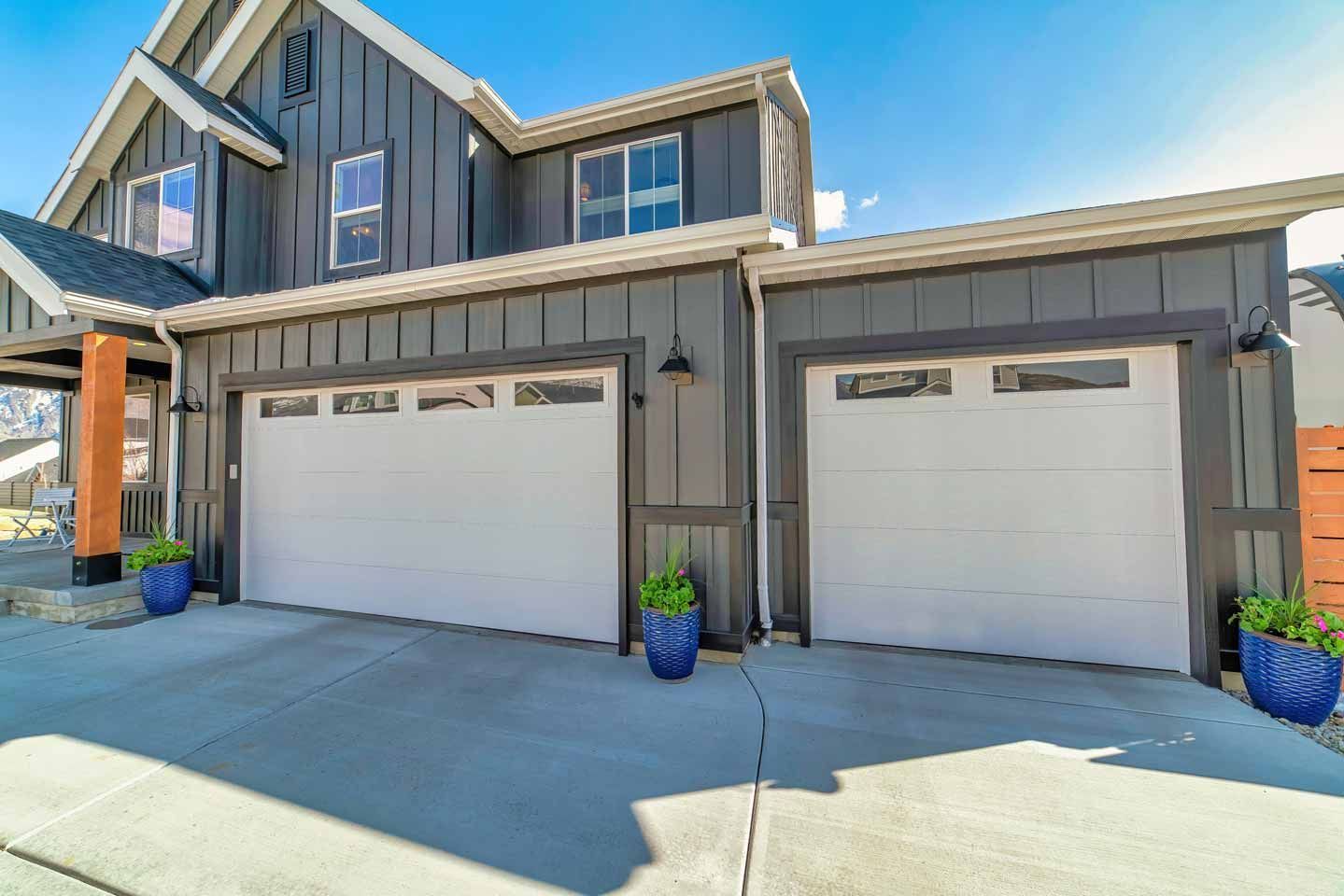 Suburban duplex with two white garage doors, gray siding, and blue potted plants by the driveway