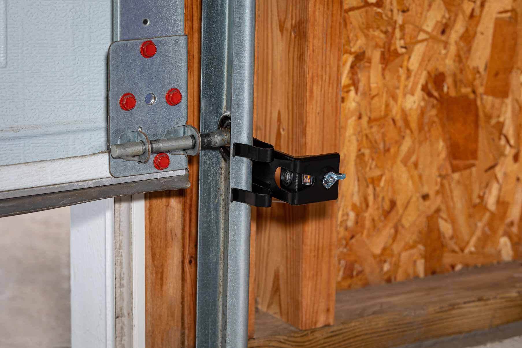 Close-up of a garage door hinge and black latch mounted on metal framing beside plywood wall.