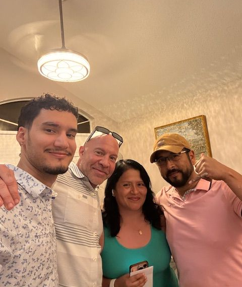 Four people smiling, posing indoors, well-lit room. Woman in teal top, man in pink shirt giving thumbs up.