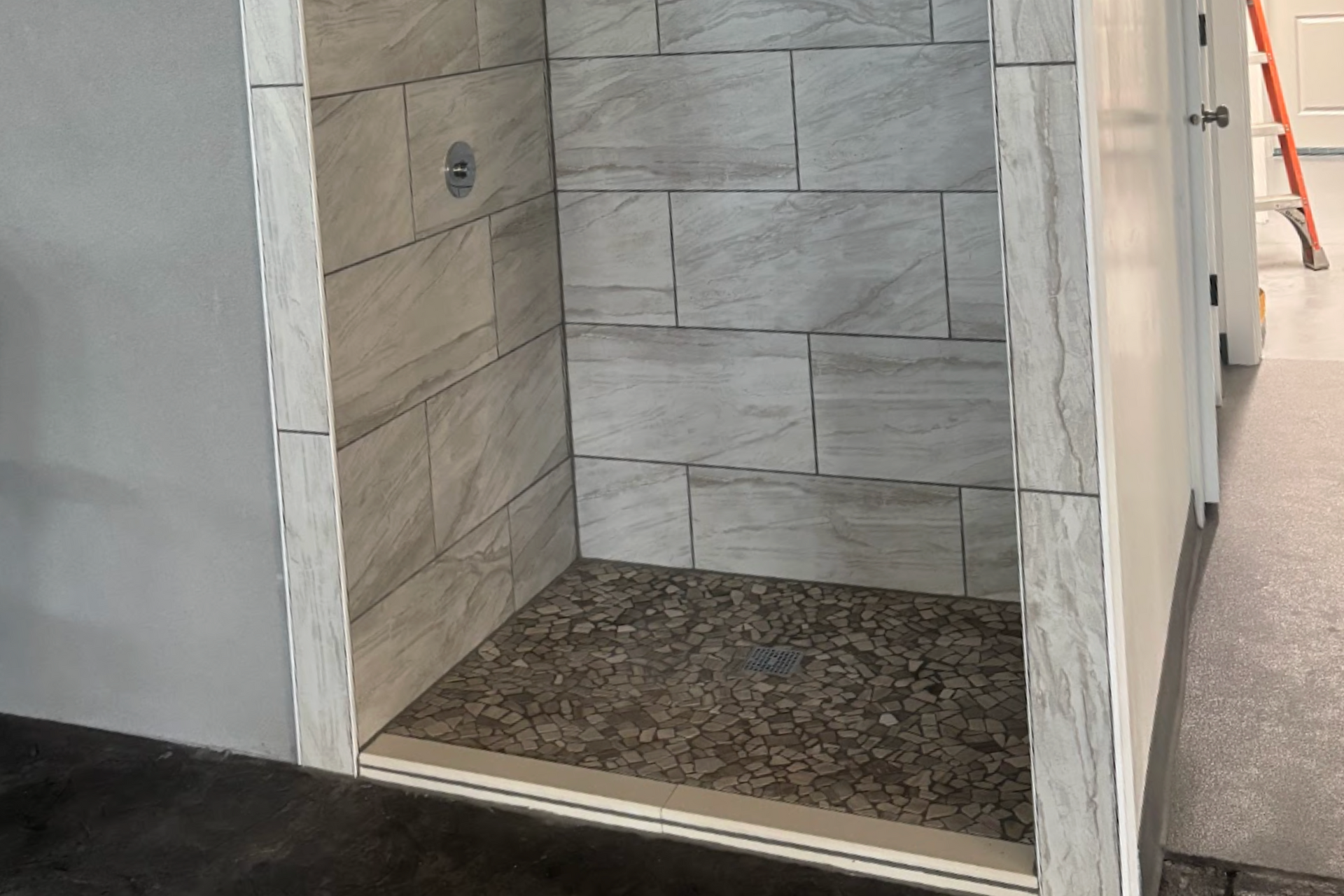 Empty tiled shower stall with a pebble floor and open doorway in a bathroom corridor