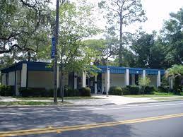 Exterior view of a one-story building with a blue roof and beige walls; set along a street with trees.