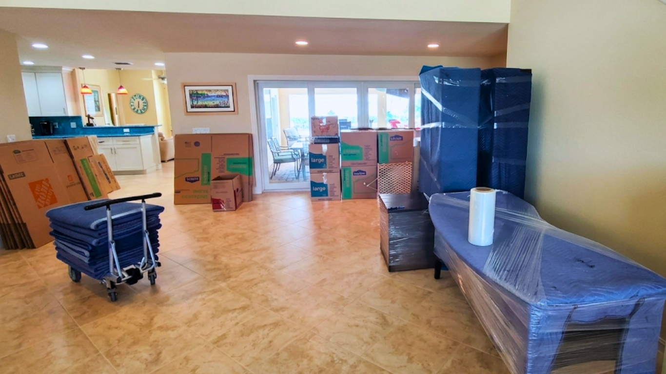 Packed room with furniture and boxes wrapped in plastic in a bright hallway or lobby.