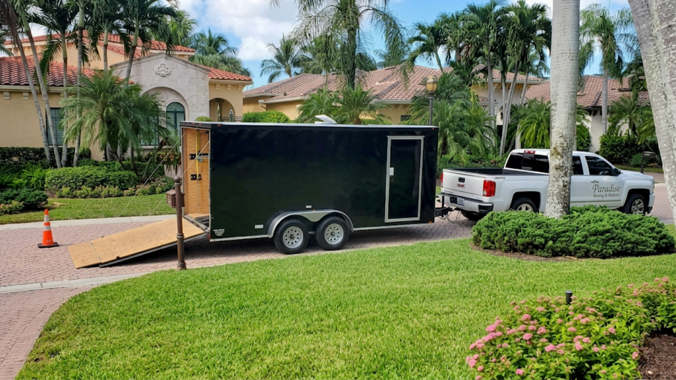 Black enclosed trailer parked on a suburban street beside a white pickup truck and palm trees