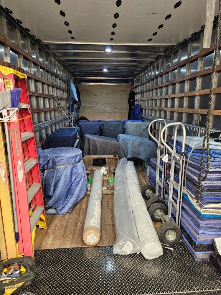 Inside a cargo van, shelves line both sides with stacked supplies and rolled rugs or mats on the floor.