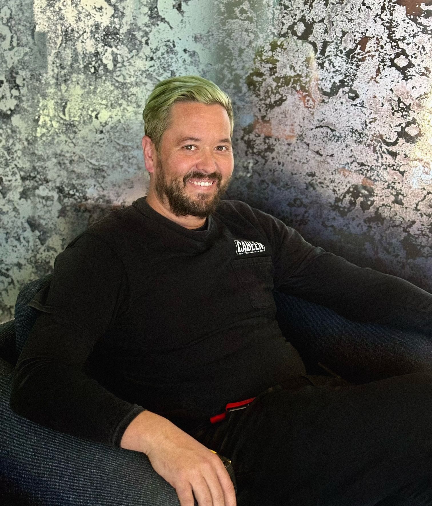 A man with a beard is sitting in a chair wearing a black shirt.