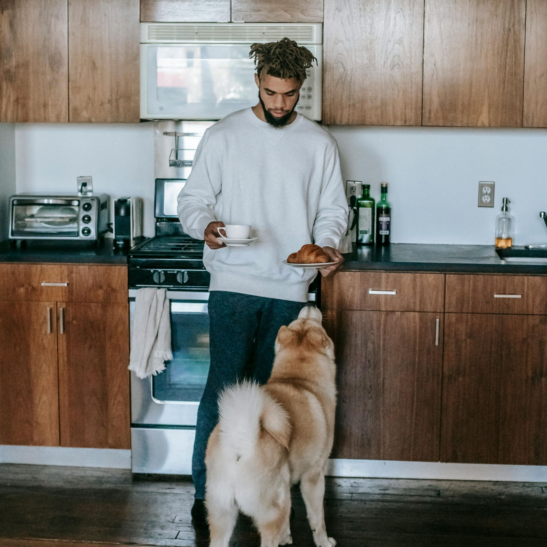 Man in a white sweater in kitchen holding food, dog looking up.