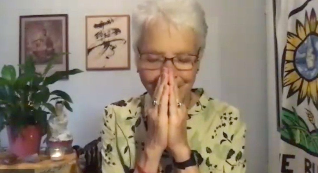 Woman with white hair and glasses, hands clasped near face, smiling. Interior with art, plants.