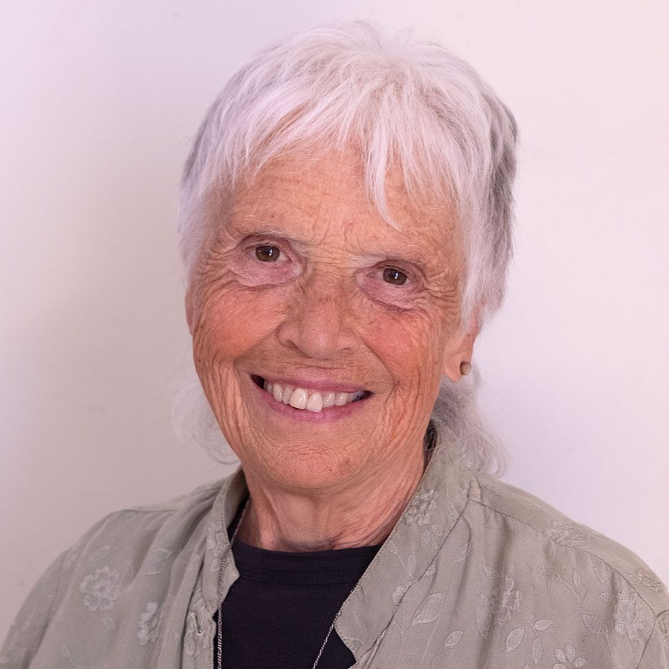 Bestsy Rose. Woman with white hair and glasses, hands clasped in front of her face, smiling. Floral shirt, artwork in background.