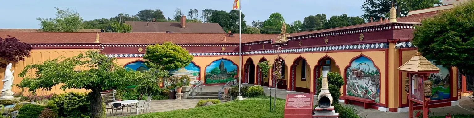 Gyuto Foundation. A courtyard with ornate, peach-colored buildings, arched doorways, and murals. Green grass and trees are in the foreground.
